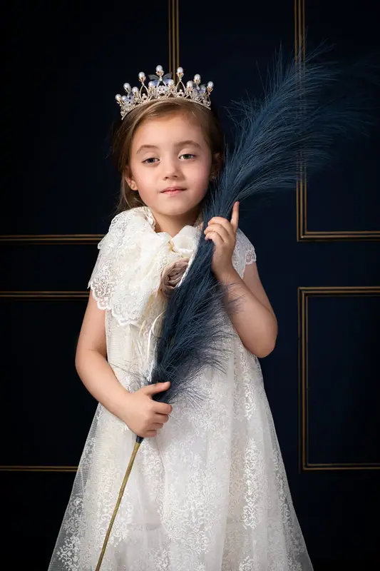 Girl in a white lace dress and crown holding a feather, dark blue backdrop