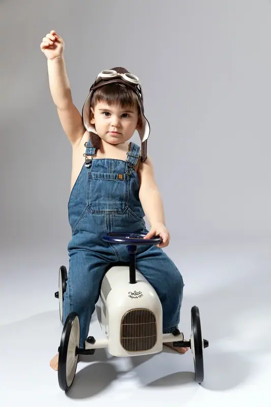 Girl in denim dungarees standing on a toy pedal car with one arm raised