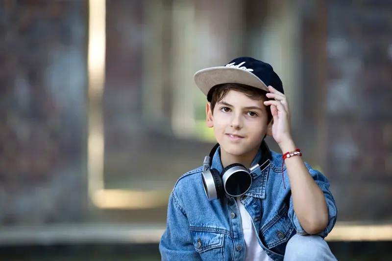 Boy in a denim jacket and cap with headphones around his neck, brick wall behind