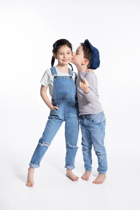 Two children in denim standing together on a white background, one kissing the other's cheek