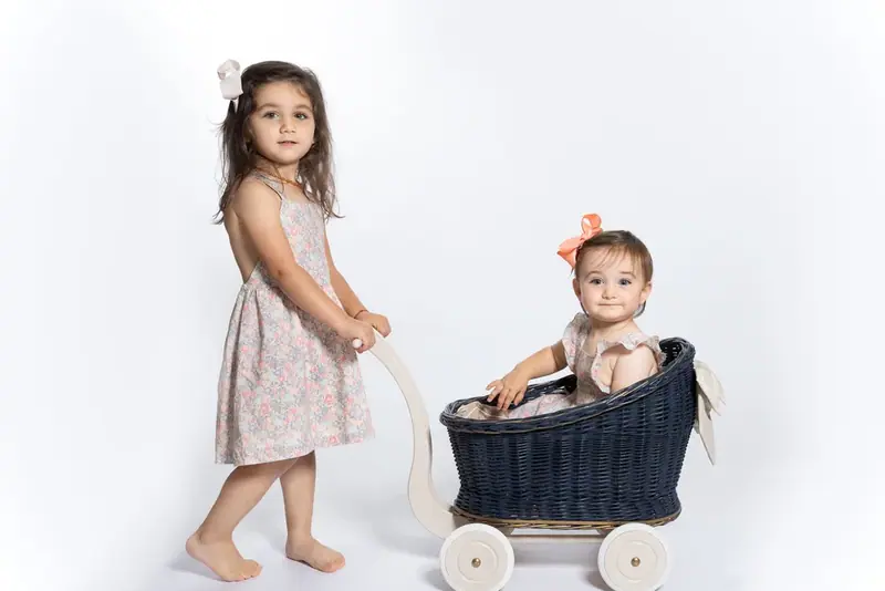 Girl in a floral dress pushing a toy pram with a baby sitting in it, white background