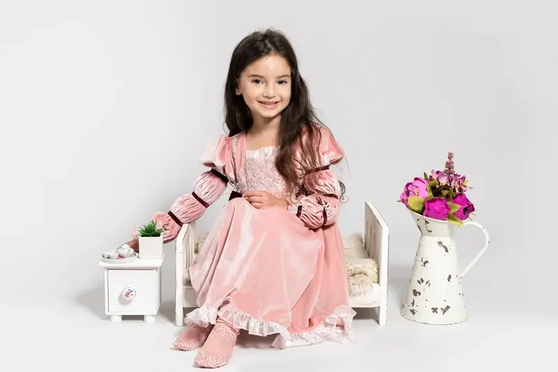 Girl in a pink dress sitting on a stool beside a jug of flowers, white background