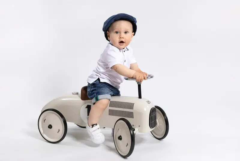 Toddler boy in a flat cap riding a toy racing car, white background