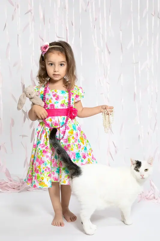 Girl in a bright floral dress standing beside a white cat, white backdrop