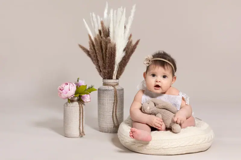 Baby sitting on a cushion beside vases of dried grasses and pink flowers