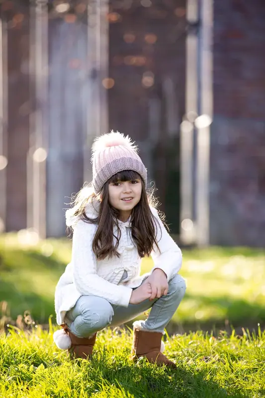Girl in a pink bobble hat crouching on the grass in front of an old brick building