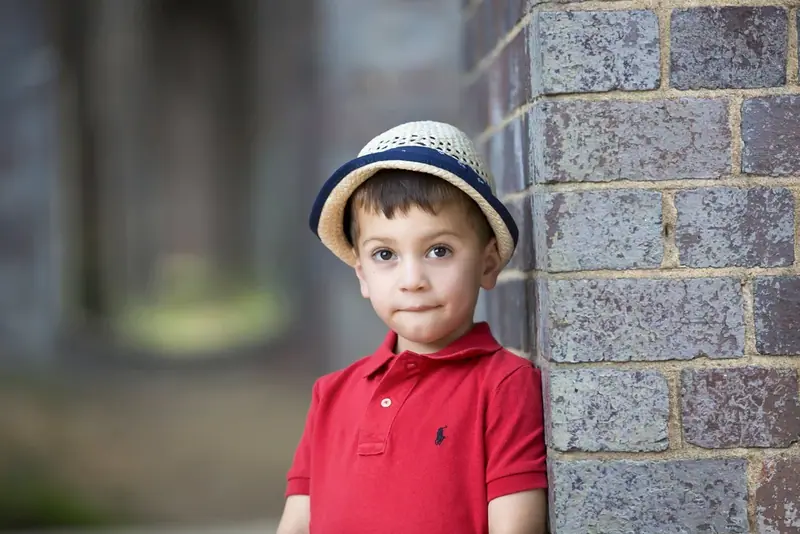 Boy in a straw hat and red polo shirt leaning against a brick wall