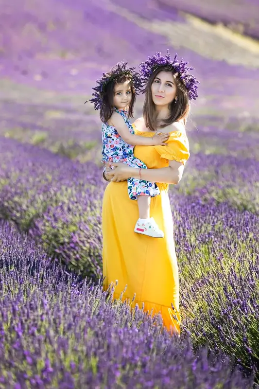 Mother in a yellow dress holding a baby in a lavender field, both wearing flower crowns