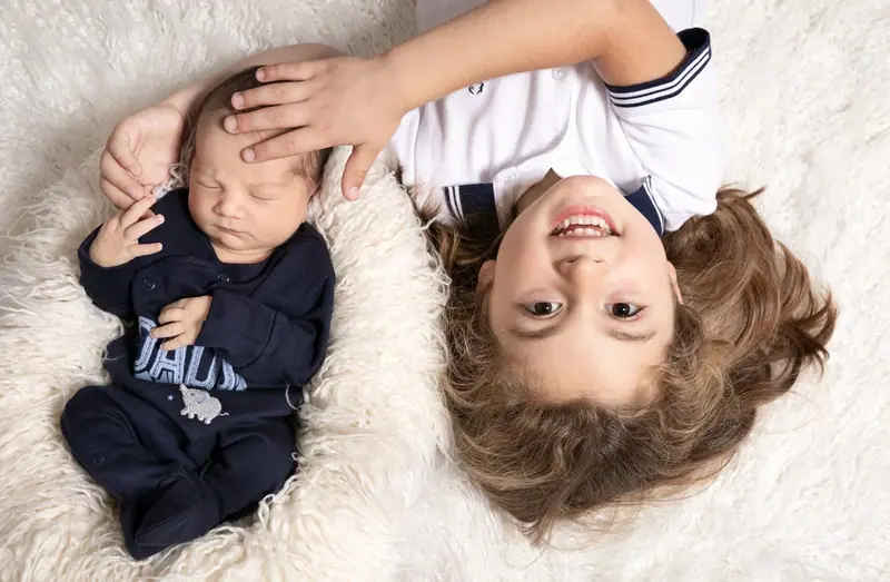 Boy lying head to head with a newborn on a white rug, seen from above