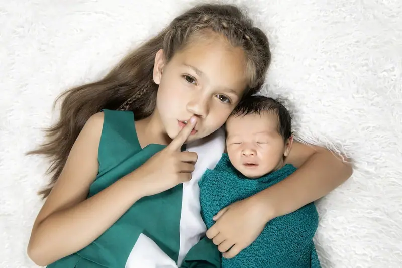 Girl in green lying beside a newborn with a finger to her lips, seen from above