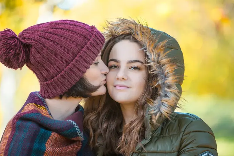 Mother in a bobble hat kissing her daughter's cheek outdoors in winter