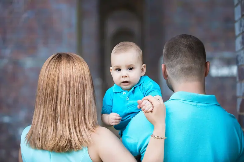 Parents in blue holding a baby, photographed from behind by a brick wall
