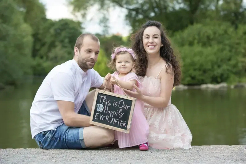 Parents and a young daughter holding a small chalkboard sign by a lake