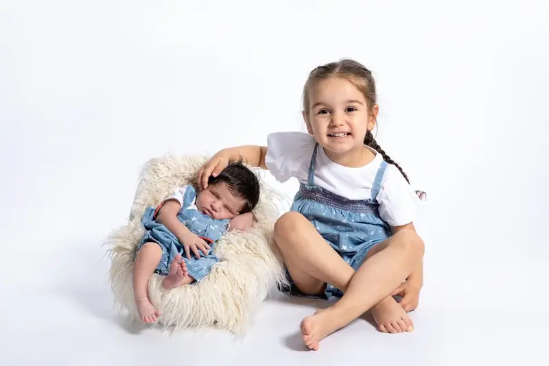 Young girl in dungarees sitting beside a newborn on a small chair