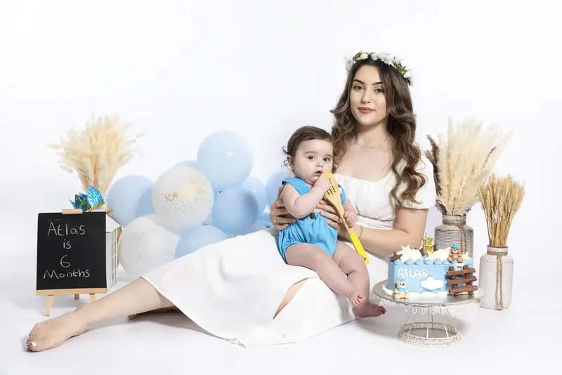 Mother in a flower crown lying beside a baby with pale blue balloons and a chalkboard sign