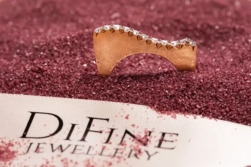 Curved diamond ring on a red glitter surface beside a jewellery brand card