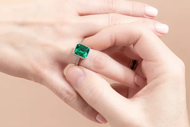 Hand holding an emerald ring, close-up on a peach background