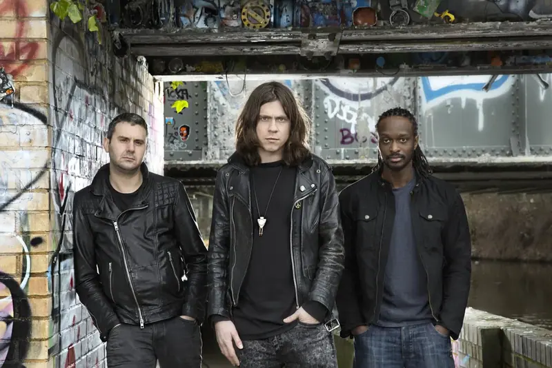 Three musicians in black leather jackets in front of a brick wall