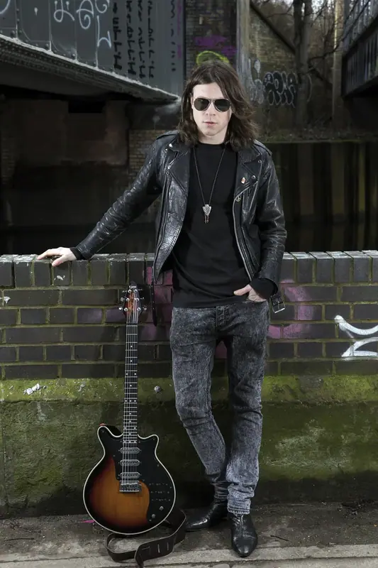Musician in sunglasses holding a guitar beside a brick wall