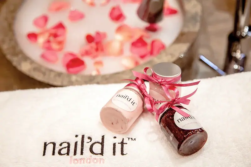 Two nail polish bottles with ribbon beside a bowl of petals and a brand logo