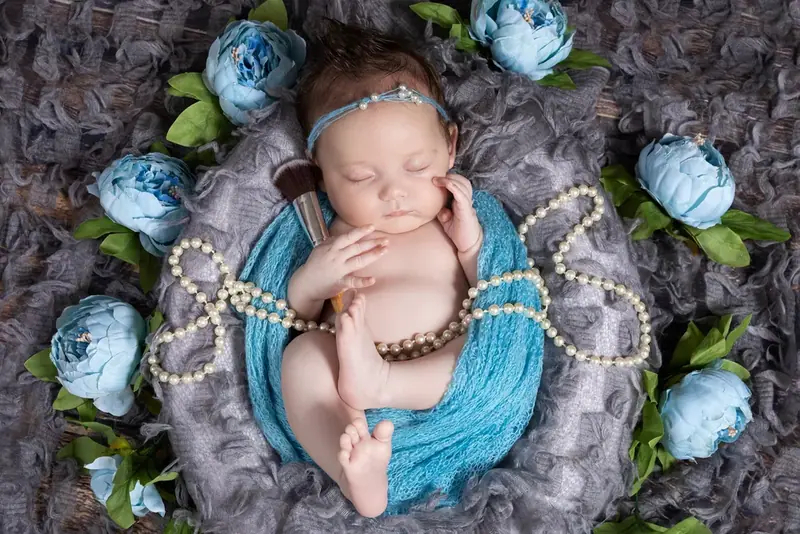 Newborn baby lying on blue and grey fabric with pale blue flowers and a string of pearls