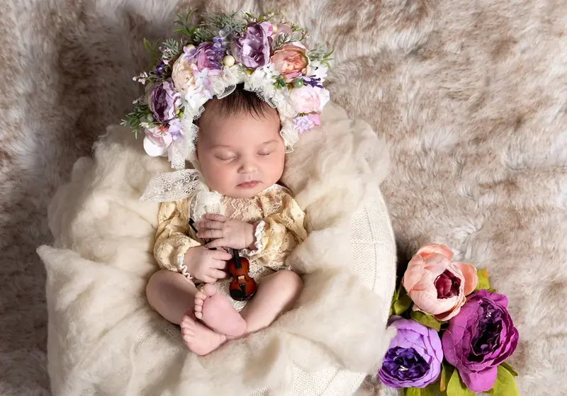 Newborn wearing a floral headband, lying on a cream fur blanket beside purple flowers