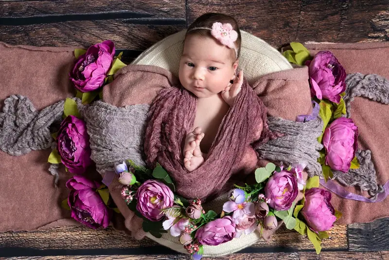 Newborn wrapped in plum fabric on a brown blanket, surrounded by pink and purple flowers