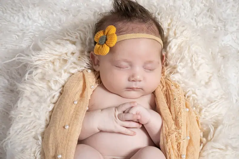 Newborn in a yellow hat lying on cream fur with a rose and pearls