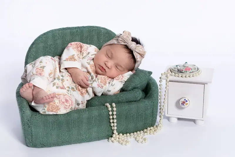 Newborn asleep in a tiny green armchair with flowers and a string of pearls