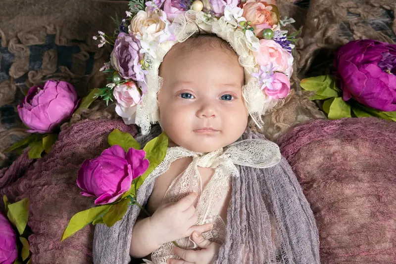 Newborn wearing a flower crown, lying on grey fur between pink and purple flowers
