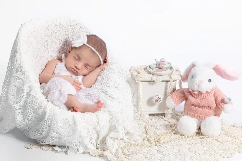 Newborn in a cream bonnet lying on white fur beside a toy rabbit