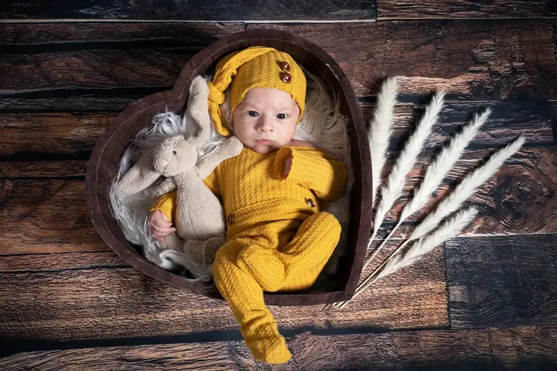Newborn in a mustard knitted outfit and hat lying in a heart-shaped wooden bowl with dried grasses