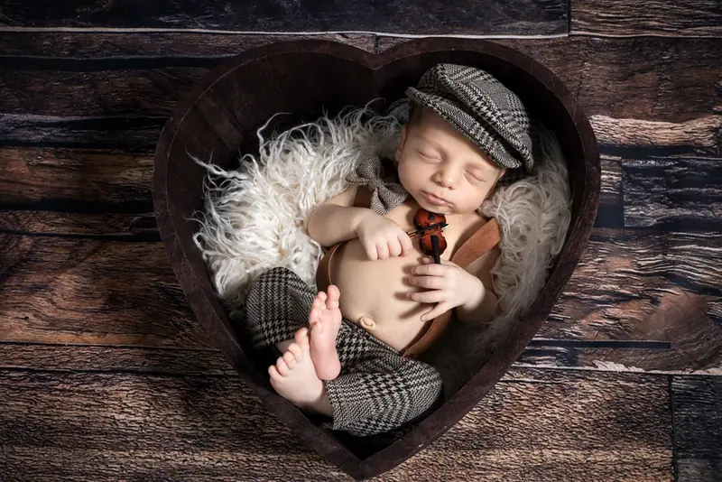 Newborn in a flat cap curled up in a heart-shaped wooden bowl on fur