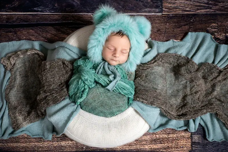 Newborn in a teal knitted hood wrapped in a green blanket on a wooden floor