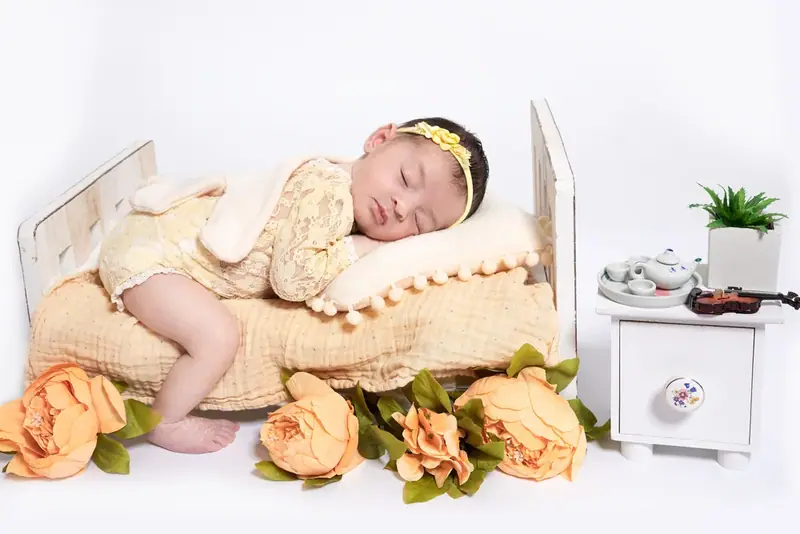 Newborn asleep in a small wooden bed with orange flowers and a tiny bedside table