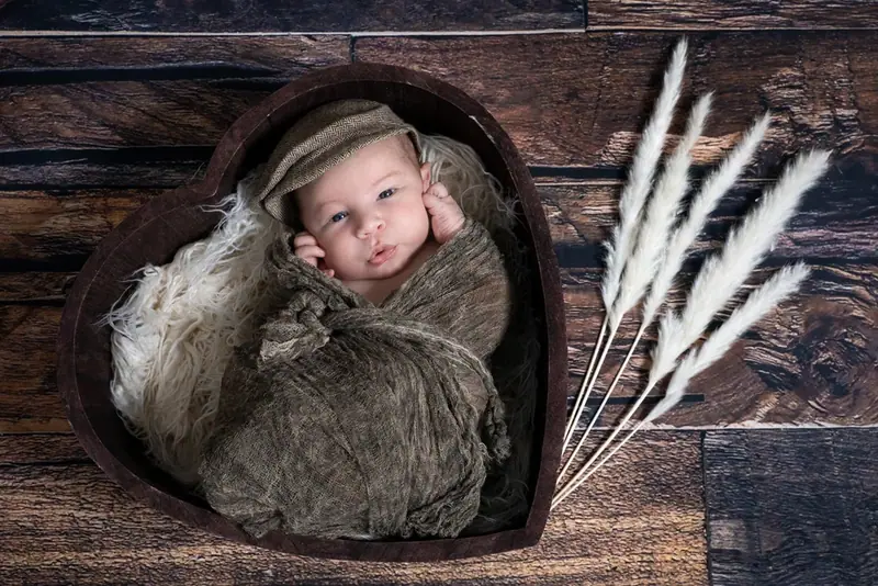 Newborn in a flat cap curled up in a heart-shaped wooden bowl on a dark wooden floor