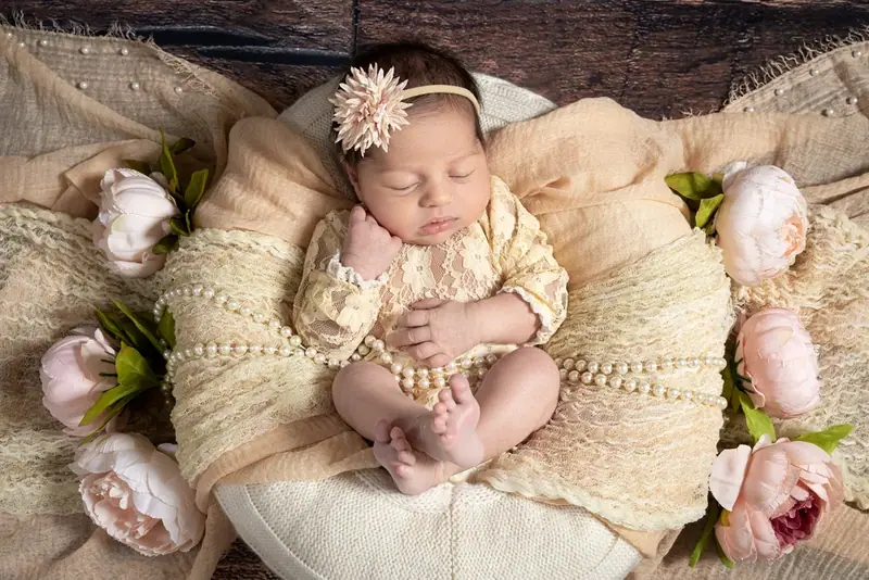 Newborn in a cream bonnet wrapped in a yellow blanket beside pink flowers