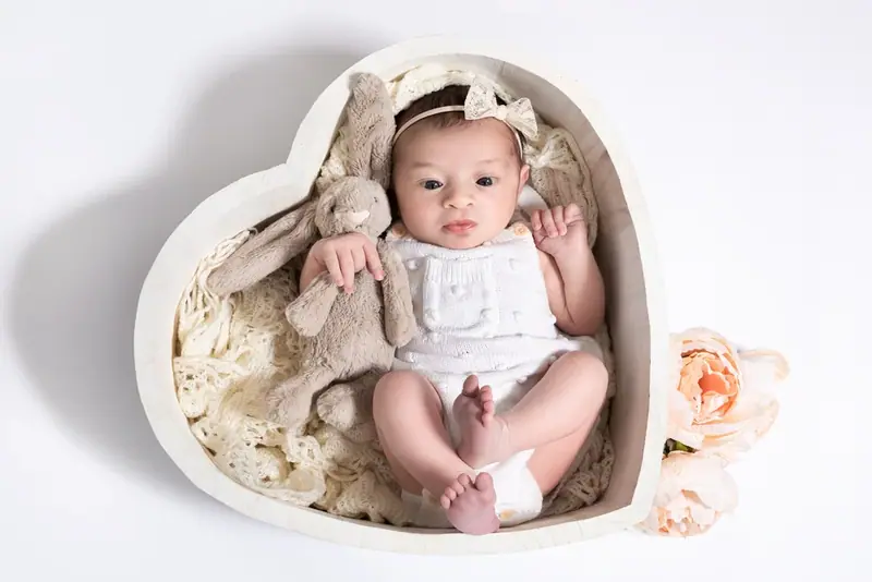 Newborn in a lace headband lying in a heart-shaped wooden bowl on white fur