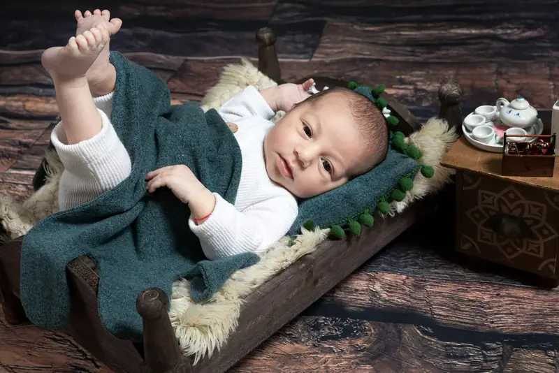 Newborn in a white outfit lying on a green blanket in a wooden crate beside a small table