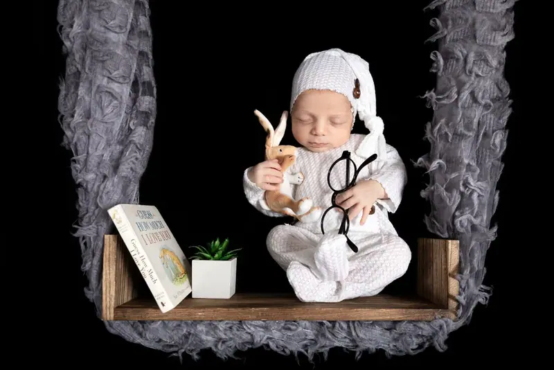 Newborn in a white outfit seated on a wooden swing with toy glasses and a toy rabbit