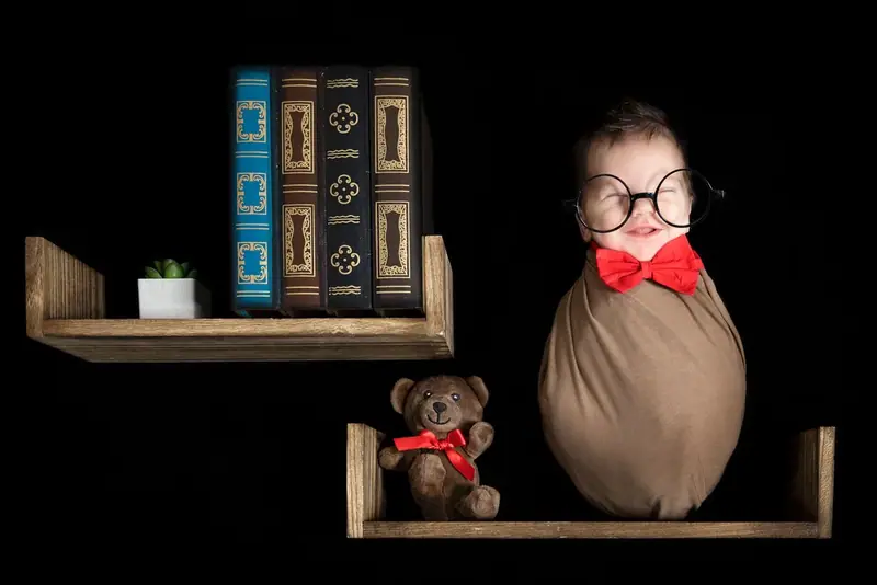 Newborn wrapped in brown with a red bow tie, posed on a wooden shelf with books and a teddy bear
