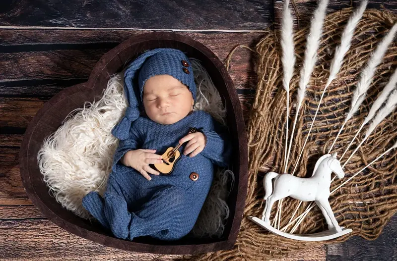 Newborn in a navy knitted outfit in a heart-shaped bowl beside a toy rocking horse