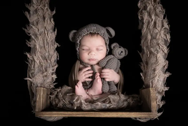 Newborn in a bear hat cuddling a teddy bear on a wooden swing