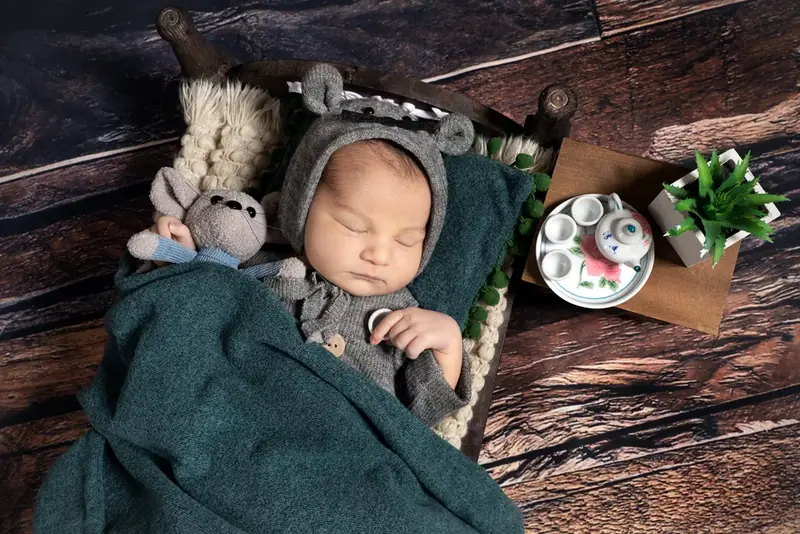 Newborn in a knitted hat wrapped in a green blanket beside a toy bear and a small plant