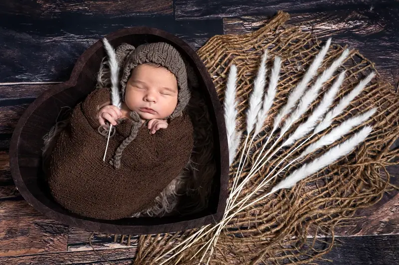 Newborn asleep in a brown wrap in a heart-shaped bowl with dried grasses