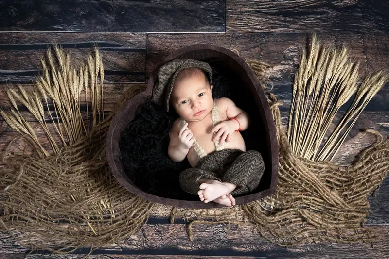 Newborn in a brown hat curled up in a heart-shaped wooden bowl with dried wheat