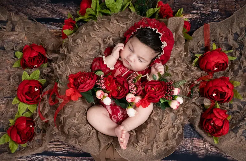 Newborn in a red outfit and red headband lying among red roses
