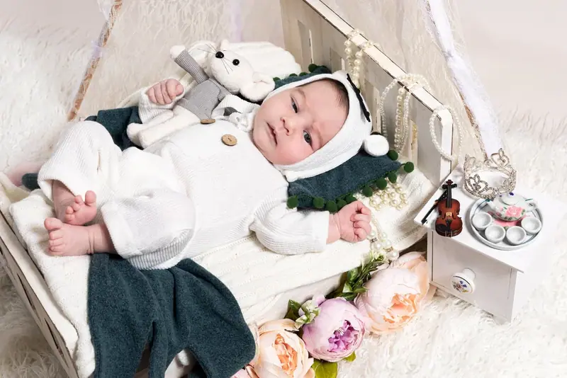 Newborn in a white outfit lying in a small wooden bed with a toy and flowers