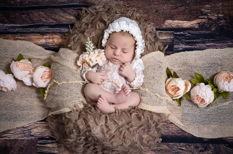 Newborn in a cream bonnet lying on a lace blanket beside orange flowers
