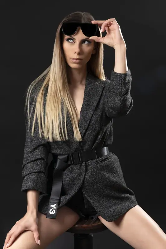 Woman in a dark belted blazer and sunglasses posing in a dark studio
